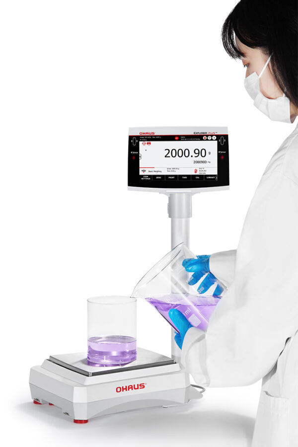 Ohaus Explorer Precision Balance viewed side on this person decanting liquid into a beaker. White background.